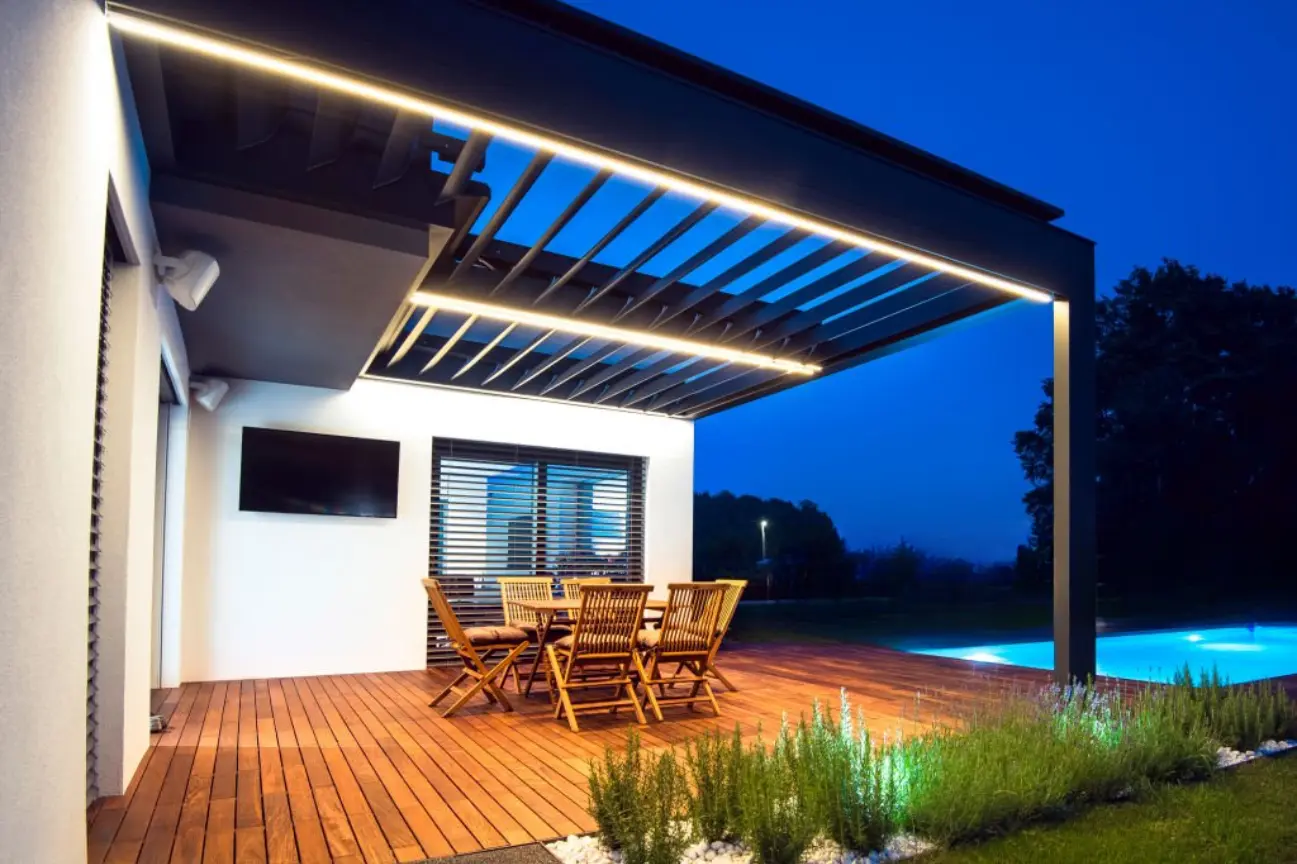 A modern louvered aluminium pergola with integrated LED lighting over a wooden deck patio at night by RHL Build HB Pergolas.