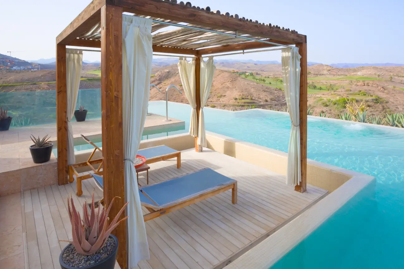 A resort-style wooden cabana pergola with white draping curtains set beside an infinity pool by RHL Build HB Pergolas.