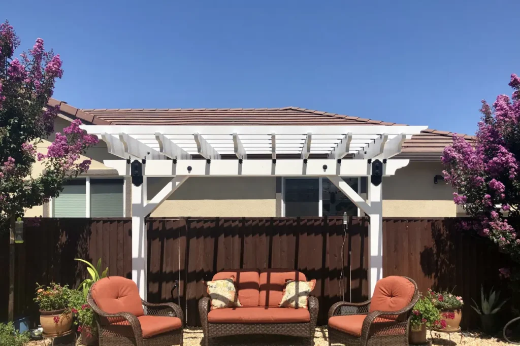 A stylish white backyard pergola with cozy orange cushioned seating framed by blooming flowers built by RHL Build HB Pergolas.